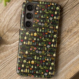May include: A black smartphone case with a botanical design featuring shelves of plants, flowers, and apothecary bottles. The case is on a wooden surface with a camera and greenery in the background.
