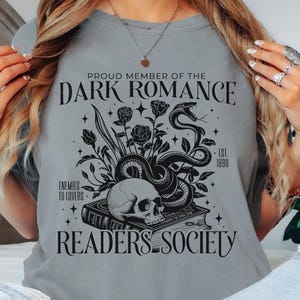 May include: A grey t-shirt with a black graphic design. The design features a skull, a snake, flowers, and a book. The text on the design reads 'Proud Member of the Dark Romance Readers Society - Est 1890 - Enemies to Lovers'.
