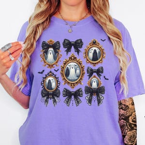May include: A violet t-shirt featuring a Halloween-themed design. The design includes framed ghost portraits, black bows, and bats. The word "Violet" is printed on the right side of the shirt.