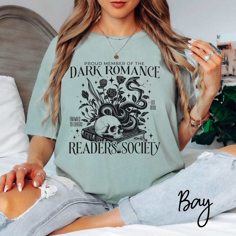 May include: A light blue t-shirt with a black graphic design featuring a skull, snake, and book. The text on the shirt reads "Proud Member of the Dark Romance Readers Society - Est. 1890 - Enemies to Lovers".