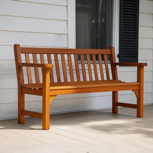 May include: A wooden garden bench with a slatted back and armrests. The bench is made of reddish-brown wood and is positioned on a light-coloured wooden deck. The background features a white wall and a window with black shutters.