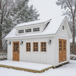 May include: A white wooden shed with a gabled roof, a dormer with three small windows, and a front deck. The structure features natural wood-toned doors with cross-bracing, multiple window panes, and black wall-mounted lanterns during a snowfall.
