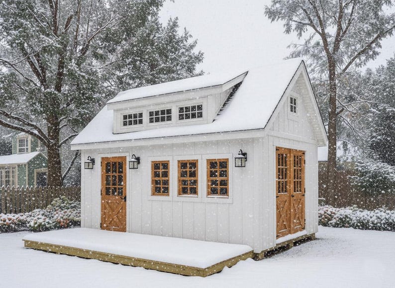May include: A white wooden shed with a gabled roof, a dormer with three small windows, and a front deck. The structure features natural wood-toned doors with cross-bracing, multiple window panes, and black wall-mounted lanterns during a snowfall.