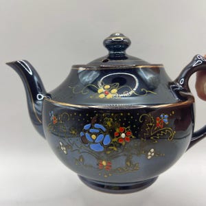 May include: A black teapot with gold trim and floral designs in blue, red, yellow, and white. The teapot has a lid with a small hole in the top.