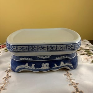 May include: A rectangular ceramic display stand featuring a white top surface and a blue and white decorative pattern along the sides. The base has a scalloped edge with intricate blue floral and geometric motifs painted on the ceramic body.