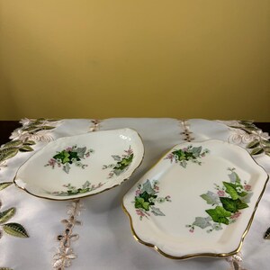 May include: Two off-white ceramic dishes with scalloped edges and gold trim. The dishes feature a botanical pattern of green ivy leaves and small pink flowers scattered across the surface. One dish is oval-shaped, and the other is rectangular.