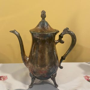 May include: An antique silver-plated coffee pot with a curved spout and ornate handle. The pot has a domed lid with a decorative finial and rests on three small feet. The metal has a tarnished appearance, showing shades of brown and blue.