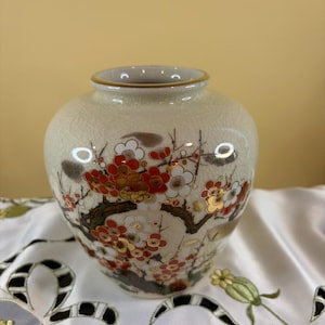 May include: A cream-colored ceramic vase with a crackle glaze finish, featuring a detailed hand-painted design of flowering plum branches in shades of red, white, and gold. The vase has a rounded body and a narrow, circular opening with a gold rim.
