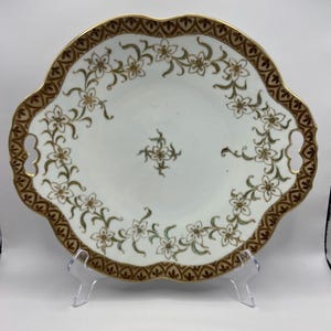 May include: A white porcelain serving plate with a scalloped edge and two handles. The plate is decorated with a gold rim and a floral pattern in green and gold.
