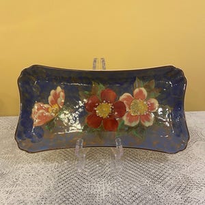 May include: Rectangular ceramic serving dish with a dark blue background and floral design. The flowers are red, pink, and yellow. The dish is supported by two clear plastic stands.