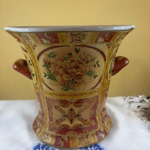 May include: A ceramic vase with a flared rim and two side handles, featuring a decorative pattern of red, gold, and cream panels. The central design displays a detailed floral motif with pink and orange flowers surrounded by intricate gold filigree.