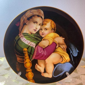 May include: A decorative plate featuring a Renaissance-style painting of a woman and child. The woman wears a green and white patterned garment and a head covering. The child is dressed in yellow. The plate has a gold rim and is displayed on a clear stand.