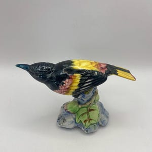 May include: A ceramic bird figurine with black, yellow, and red paint, perched on a green and blue leaf base.