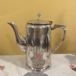 May include: A tall, polished silver coffee pot with a lid and a curved spout. The pot has a handle and sits on a round base. The reflective surface mirrors the surrounding environment, creating a unique visual effect. A classic piece for serving beverages.