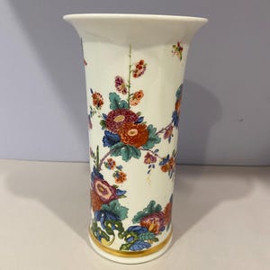 May include: A white ceramic vase with a flared top and a gold band at the base. The vase is adorned with a colorful floral pattern featuring red, orange, blue, and green flowers and foliage. The vase is approximately 8 inches tall.