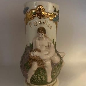 May include: A cream-colored ceramic vase with a relief sculpture of two figures. The word "Paradise" is written in gold lettering. The vase has gold accents and a detailed base.