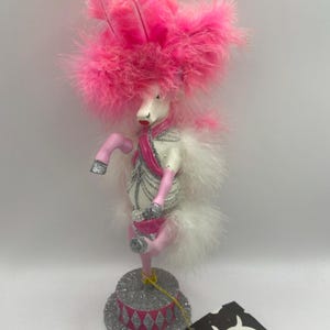 May include: A whimsical, decorative figurine of a white horse with a pink feathered hat and accents. The horse stands on a glittery silver base with a pink and silver diamond pattern. A tag is attached.