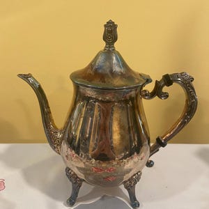 May include: A vintage silver teapot with ornate detailing. The teapot features a curved spout, a decorative handle, and rests on three small feet. The lid is topped with a decorative finial. The teapot is reflective, showing the surrounding environment.