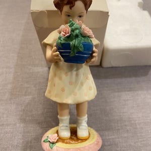 May include: A porcelain figurine of a young girl holding a blue pot of pink roses. She wears a yellow dress with peach-colored polka dots, white socks, and white shoes. The figurine stands on a round, pink and yellow base with a rose.