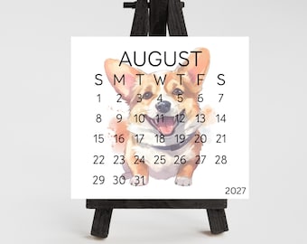 May include: A square calendar page for August 2027 featuring a watercolor illustration of a tan and white Corgi dog. The calendar displays the month, days of the week, and dates. It is displayed on a small black wooden easel. Dimensions are 3.25" W x 3" H.