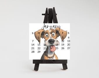 May include: A square January 2024 calendar card featuring a cartoon dog with brown and white fur, large eyes, and a pink tongue sticking out. The calendar is displayed on a small black wooden easel stand against a plain white background.