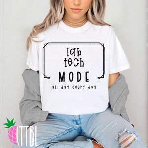 May include: White t-shirt with the text "lab tech MODE all day every day" in a black rectangular frame. The model is wearing blue jeans and a gray cardigan.