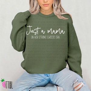 May include: An olive green sweatshirt featuring the white text "Just a mama" and "In her string cheese era". This casual, long-sleeved top is suitable for everyday wear and is a comfortable garment.