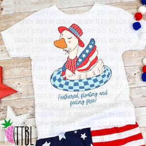 Può includere: T-shirt bianca con un'oca dei cartoni animati con cappello e gilet patriottici, che galleggia su un salvagente a scacchi blu e bianchi. Il testo "Feathered, floating and feeling free!" è sotto.