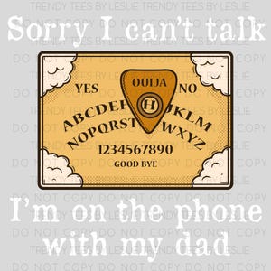 May include: A tan Ouija board with black lettering and a planchette. The board features the alphabet, numbers, and the words "Yes," "No," and "Good Bye." The text "Sorry I can't talk, I'm on the phone with my dad" is also present.