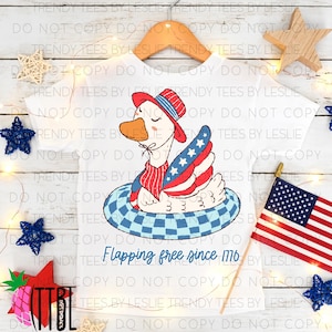 Può includere: T-shirt bianco con un'oca dei cartoni animati con cappello e sciarpa patriottici, che galleggia su un salvagente a scacchi blu e bianchi. L'oca ha ali rosse, bianche e blu con stelle. Testo: "Flapping free since 1776."