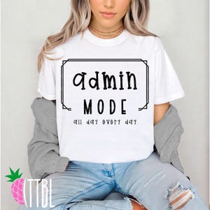 May include: White t-shirt with the words "Admin MODE" in a black, bold font, framed by a black rectangle. Below the text is "all day every day" in a smaller font. The person is wearing blue jeans.
