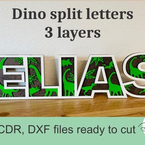 Pode incluir: Placa de madeira com o nome "ELIAS" com tema de dinossauro. As letras são brancas com recortes de dinossauros verdes e um fundo marrom. O texto "Dino split letters 3 layers" está acima.
