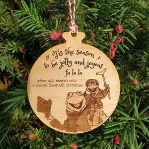 May include: Wooden Christmas ornament with a festive design. The ornament features a laser-etched image of two Muppet characters and the text "Tis the season to be jolly and joyous fa la la". It hangs from a red and white striped string.