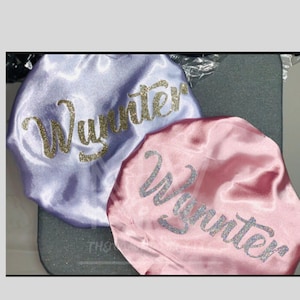 May include: Two satin bonnets, one lavender and one pink, are shown. The word "Wynnter" is written in glittery script across each bonnet. The bonnets are resting on a grey surface.