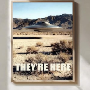 May include: A framed wall art print featuring a metallic, disc-shaped unidentified flying object hovering above a dry, cracked desert landscape with sparse shrubs and distant mountains. The text "THEY'RE HERE" is printed in white capital letters.