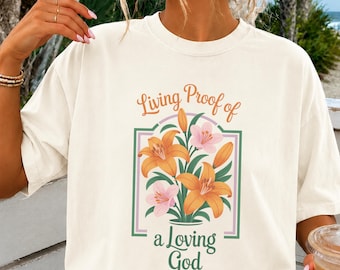May include: A cream-colored short-sleeve t-shirt featuring a graphic of orange and pink lilies in a vase. The text "Living Proof of a Loving God" is printed in orange and green script and serif fonts, framed by a thin green and purple border.