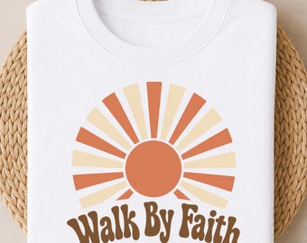 May include: A white t-shirt featuring a graphic of a sun with alternating orange and cream-colored rays. Below the sun, the text "Walk By Faith" is printed in a dark brown, rounded, retro-style font. The design is centered on the chest area.
