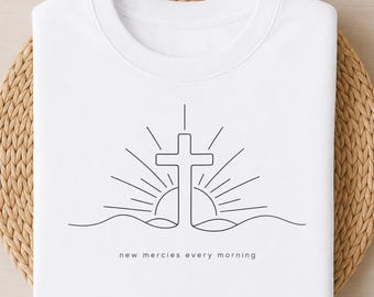 May include: A white crew neck t-shirt featuring a black line art graphic of a cross centered above a rising sun and a wavy horizon line. Below the graphic, the text reads new mercies every morning in a simple, lowercase, sans-serif font.
