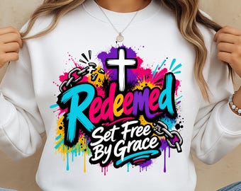 May include: A white crewneck sweatshirt featuring a colorful graphic print with a white cross, broken metal chains, and the text "Redeemed Set Free By Grace" in a bold, stylized font surrounded by vibrant paint splatters in blue, pink, and yellow.