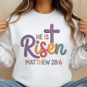 May include: White sweatshirt with the embroidered text "He Is Risen" in colorful, textured lettering, along with a cross and the Bible verse Matthew 28:6. The design features purple, yellow, green, and pink hues.