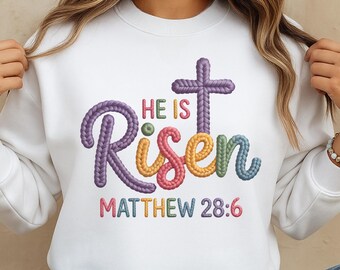 May include: White sweatshirt with the embroidered text "He Is Risen" in colorful, textured lettering, along with a cross and the Bible verse Matthew 28:6. The design features purple, yellow, green, and pink hues.