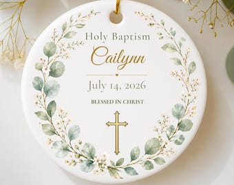 May include: A round white ceramic ornament featuring a green eucalyptus wreath design. The text reads Holy Baptism, Cailynn, July 14, 2026, and BLESSED IN CHRIST. A gold cross is centered at the bottom, and a gold string is attached at the top.