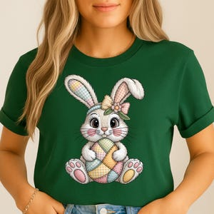 Op de afbeelding: Groen t-shirt met een cartoon konijn dat een kleurrijk paasei vasthoudt. Het konijn heeft een strik, een hoofdband en oren met patronen. Het ei is een patchwork van pastelkleuren. Het shirt is effen groen.