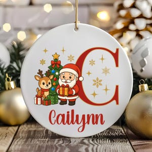 May include: White ceramic Christmas ornament with a cartoon Santa Claus, reindeer, and Christmas tree design. The ornament features the letter "C" in red and the name "Cailynn" in red script. Gold stars and snowflakes decorate the background.