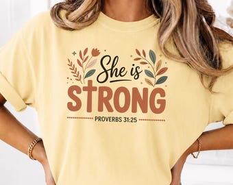 May include: A pale yellow short-sleeve t-shirt featuring a graphic design with the text "She is STRONG" and "PROVERBS 31:25". The design includes a cross integrated into the letter T and stylized botanical elements in shades of brown, green, and tan.