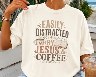 May include: An off-white short-sleeve t-shirt featuring the text "EASILY DISTRACTED BY JESUS & COFFEE" in brown and tan lettering. The graphic includes a small cross, a steaming coffee mug with a heart, and an open book illustration.