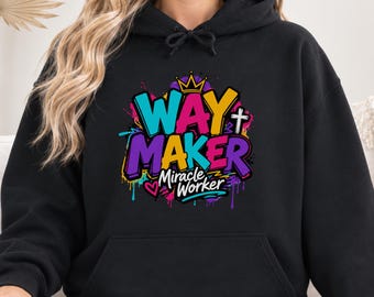 May include: A black hooded sweatshirt featuring a colorful graphic print on the chest. The design includes the text "WAY MAKER" in large, vibrant letters, "Miracle Worker" below it, a small crown, a cross, and a heart, all set against paint splatters.