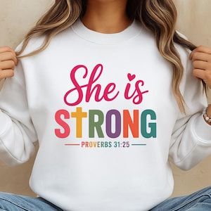 May include: White sweatshirt with the text "She is STRONG" in pink, orange, green, and blue. The text includes a heart and the Bible verse Proverbs 31:25.