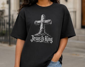 May include: A black short-sleeve t-shirt featuring a white graphic of a textured wooden cross with the text Jesus Is King written in a stylized gothic font underneath. The design has a distressed appearance with fine lines and shading.
