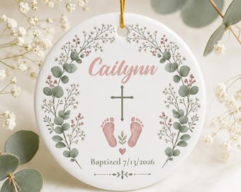 May include: A round white ceramic ornament featuring a floral wreath of green eucalyptus and small pink flowers. The center displays the name Cailynn in pink script, a dark green cross, pink baby footprints, and the text Baptized 7/13/2026.